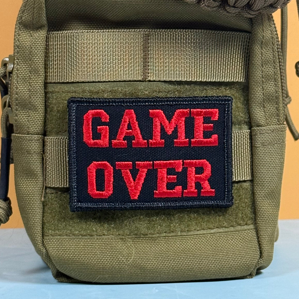 An olive green tactical pouch with a black rectangular patch featuring the words 'GAME OVER' in bold red embroidered letters, set against a solid orange background.