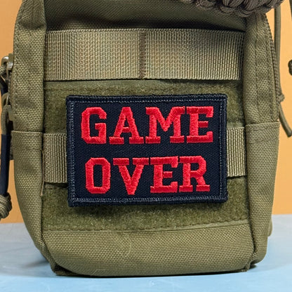An olive green tactical pouch with a black rectangular patch featuring the words 'GAME OVER' in bold red embroidered letters, set against a solid orange background.