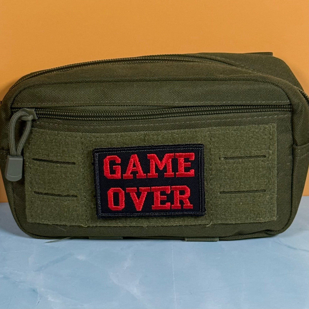 An olive green tactical pouch with a black rectangular patch featuring the red embroidered text 'GAME OVER' on the front, set against a solid orange background.
