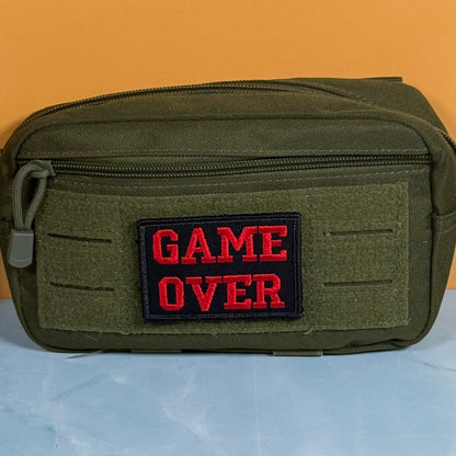 An olive green tactical pouch with a black rectangular patch featuring the red embroidered text 'GAME OVER' on the front, set against a solid orange background.