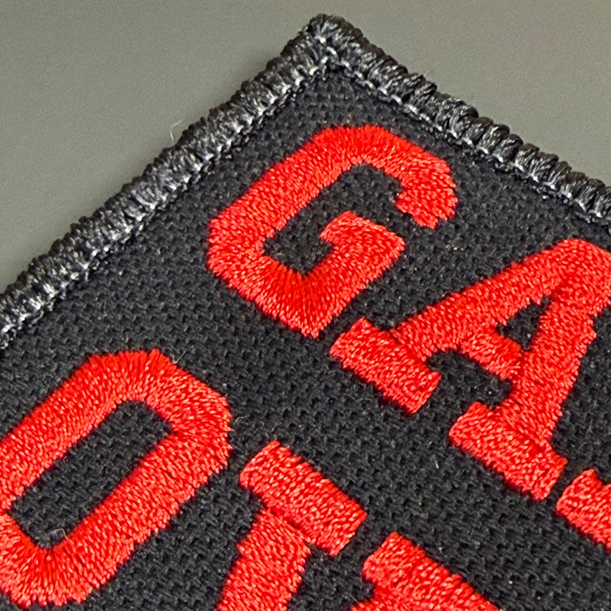 Close-up of an embroidered patch showing bold red letters on a black fabric background with a stitched border.