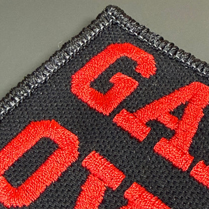 Close-up of an embroidered patch showing bold red letters on a black fabric background with a stitched border.