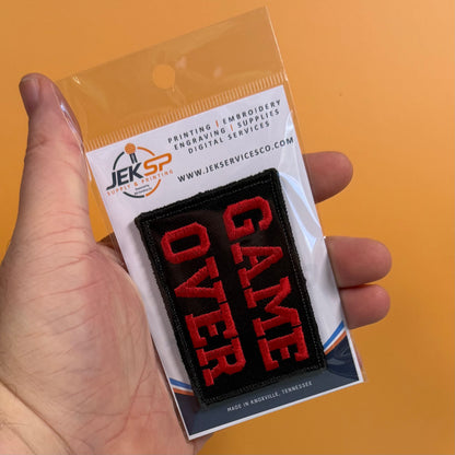 A hand holds a packaged embroidered patch that reads “GAME OVER” in red letters on a black background, attached to a white backing card with the JEKSP logo and website text, against an orange background.