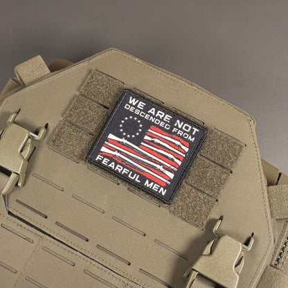 A tactical morale patch with a black background and an American flag design featuring red and white stripes and white stars. The patch reads 'WE ARE NOT DESCENDED FROM FEARFUL MEN' and is shown attached to tan tactical gear with velcro and plastic buckles.