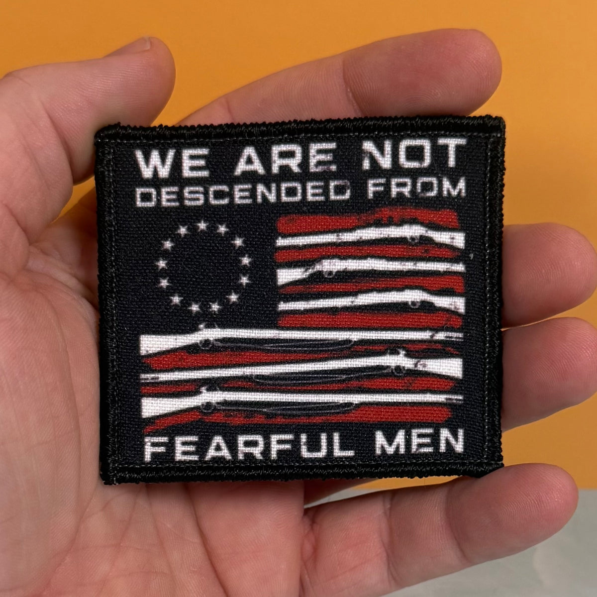 A hand holding a square embroidered patch with a black border and a distressed American flag design in red, white, and black. The patch reads 'WE ARE NOT DESCENDED FROM FEARFUL MEN' in white text.