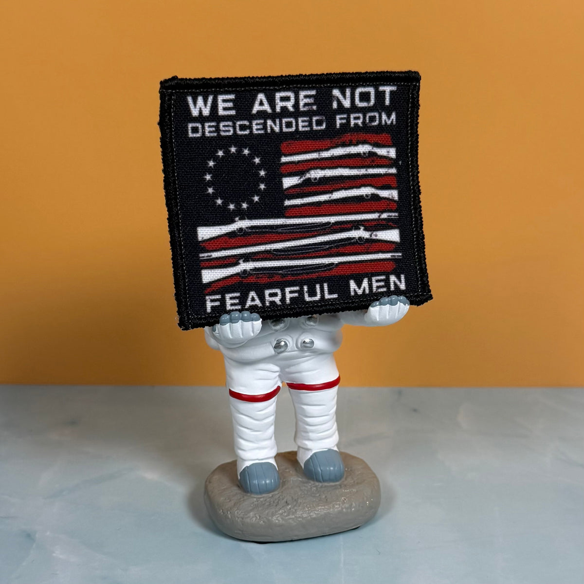 A square embroidered patch with a black background and a stylized American flag made from rifles, with the text 'WE ARE NOT DESCENDED FROM FEARFUL MEN' in white. The patch is held by a small astronaut figurine standing on a gray rock base against a warm orange background.