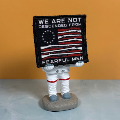A square embroidered patch with a black background and a stylized American flag made from rifles, with the text 'WE ARE NOT DESCENDED FROM FEARFUL MEN' in white. The patch is held by a small astronaut figurine standing on a gray rock base against a warm orange background.