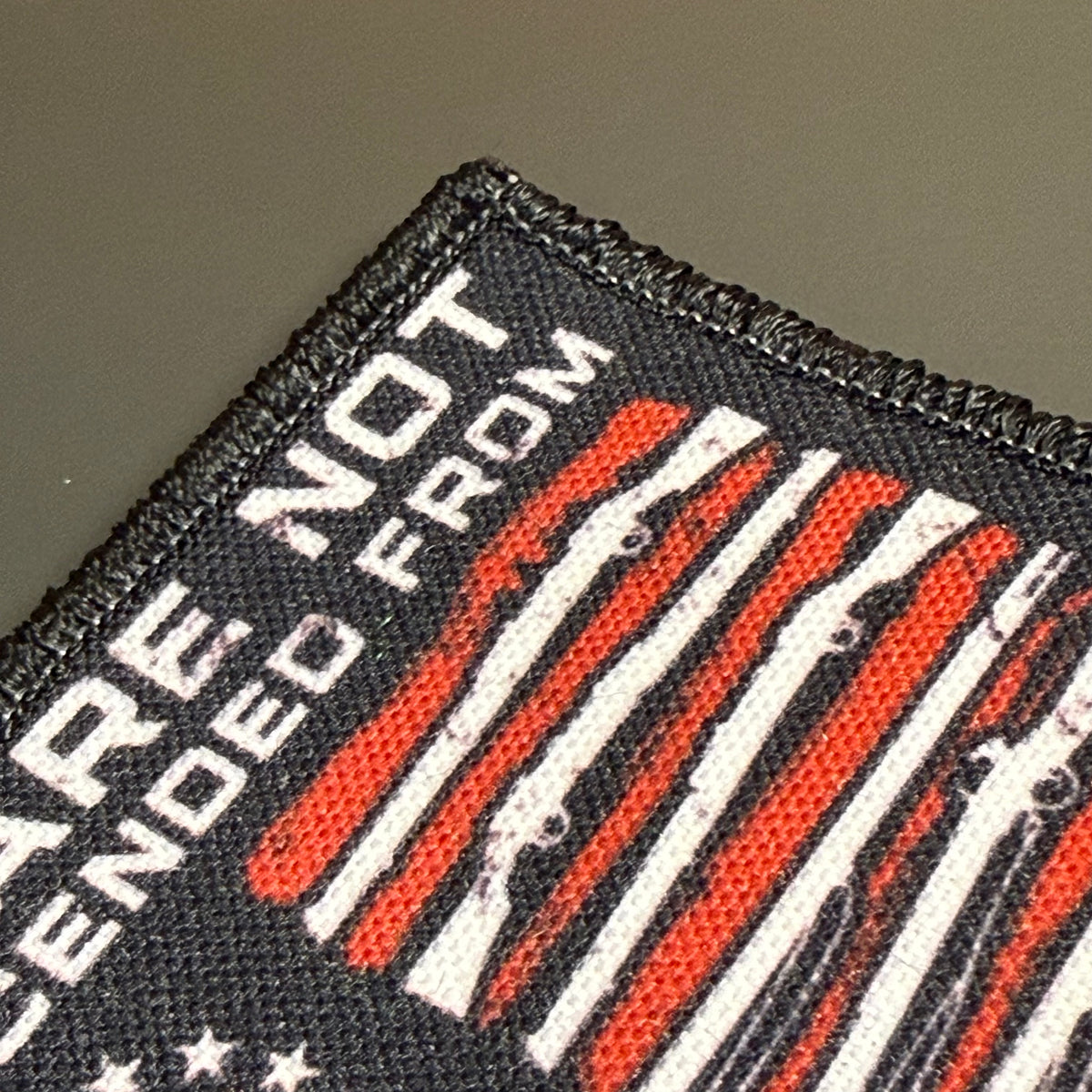 Close-up of a black embroidered patch with white text reading 'ARE NOT GENDED FROM' and a distressed red and white striped flag design, shown on a neutral gray background.