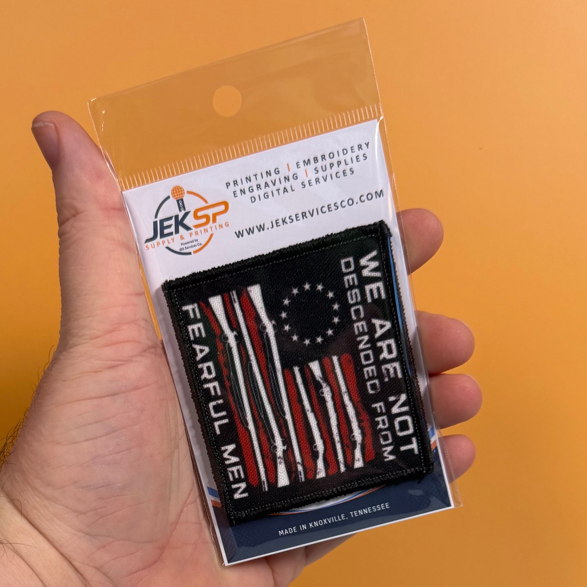 A hand holds a packaged embroidered patch featuring a distressed American flag design with red, white, and black stripes and the text 'WE ARE NOT DESCENDED FROM FEARFUL MEN.' The packaging card shows the Jeksp Digital Services brand and website, with a solid orange background behind the hand and package.