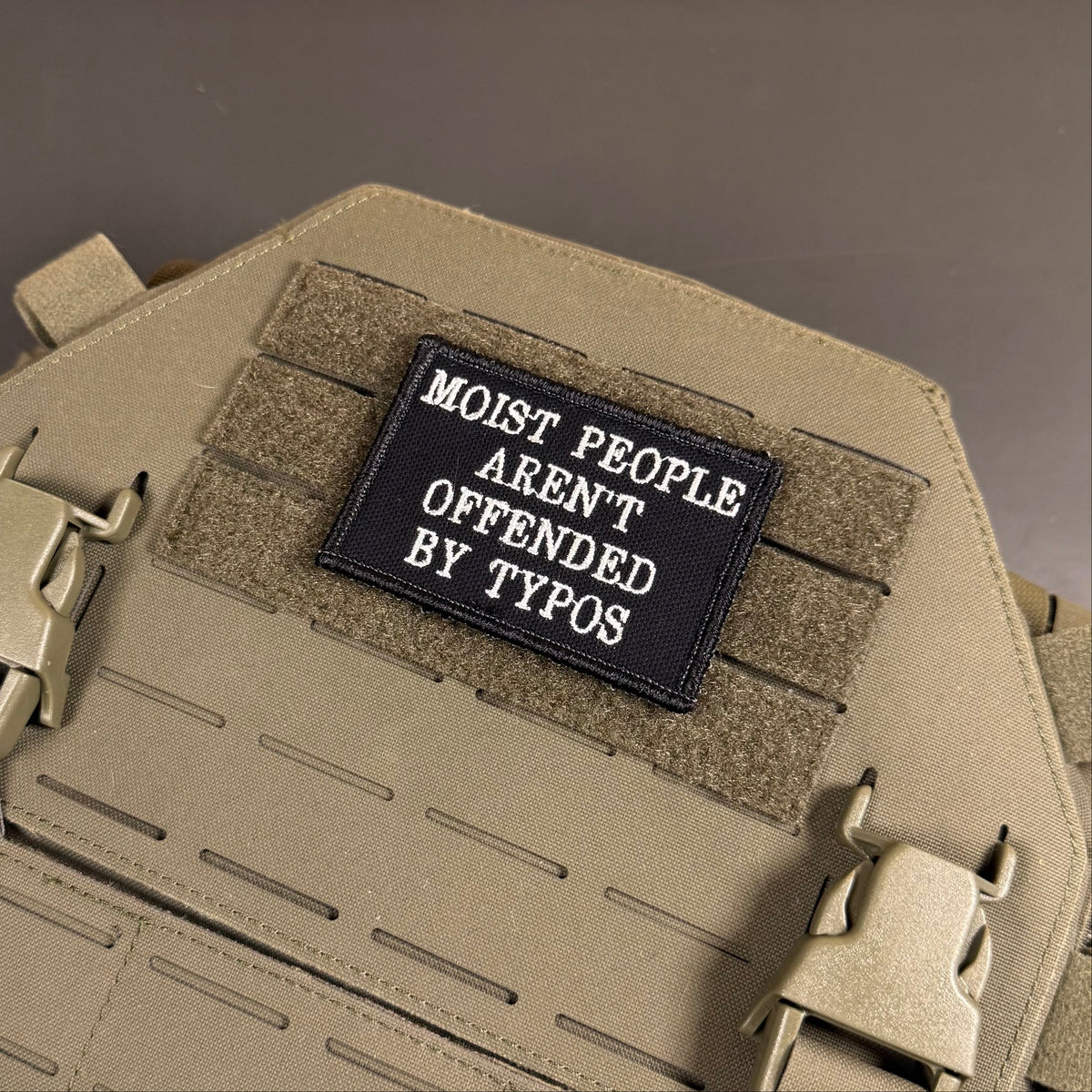 Moist People Aren’t Offended by Typos embroidered morale patch: tactical vest or plate carrier in tan/khaki fabric, featuring a black rectangular embroidered patch with white text that reads 'MOIST PEOPLE AREN'T OFFENDED BY.