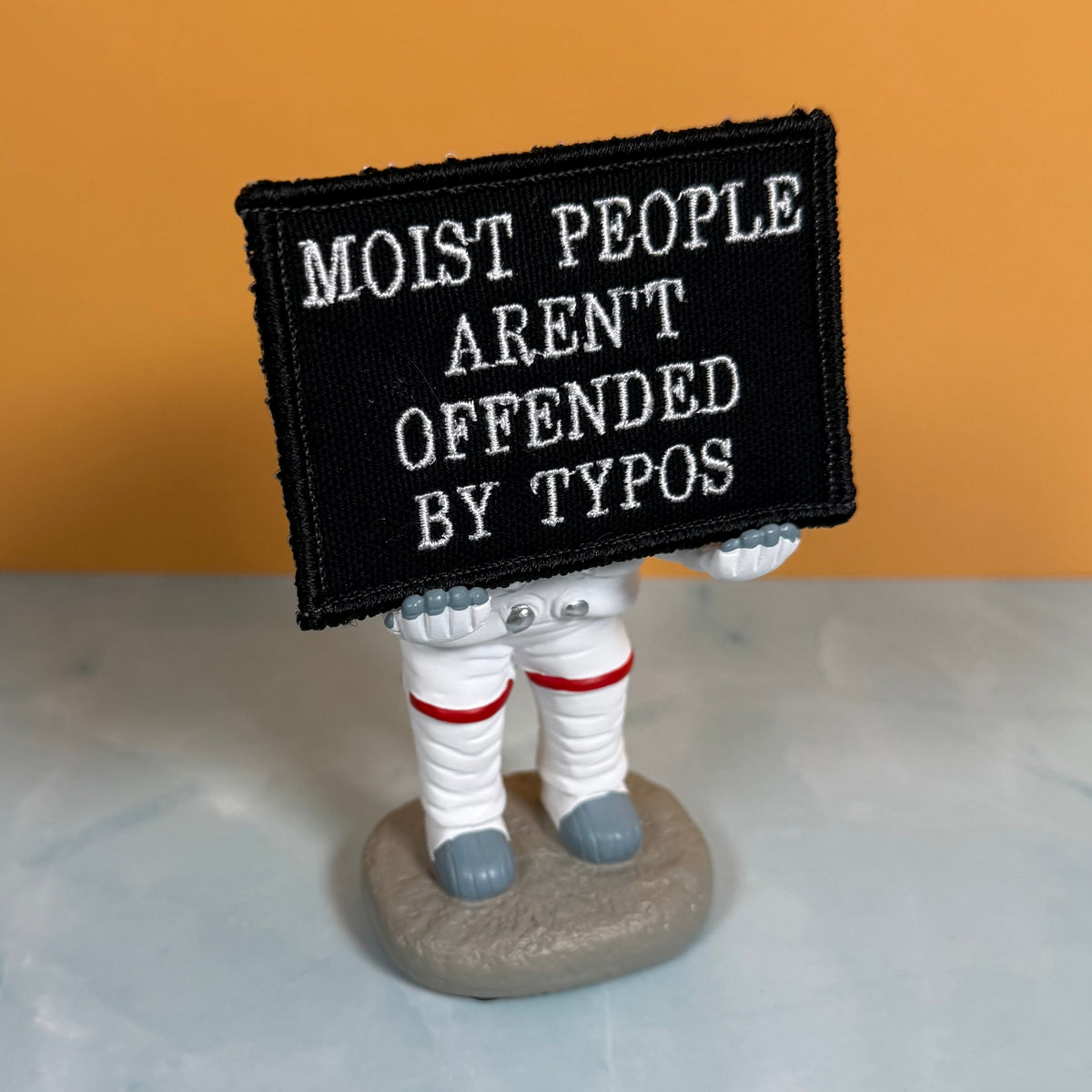Moist People Aren’t Offended by Typos embroidered morale patch: small astronaut figurine holding a black embroidered patch with white text that reads, 'MOIST PEOPLE AREN'T OFFENDED BY TYPOS.' The figurine stands on a gray.
