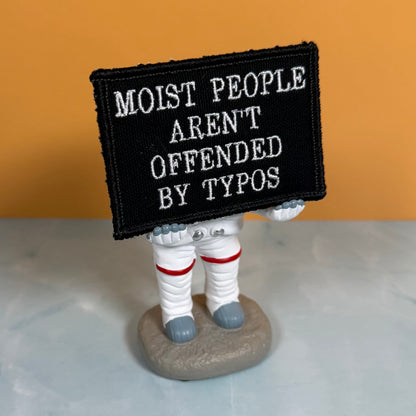 Moist People Aren’t Offended by Typos embroidered morale patch: small astronaut figurine holding a black embroidered patch with white text that reads, 'MOIST PEOPLE AREN'T OFFENDED BY TYPOS.' The figurine stands on a gray.
