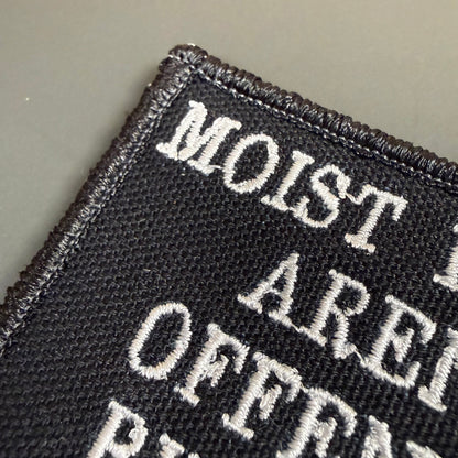 Moist People Aren’t Offended by Typos embroidered morale patch: black embroidered patch with white text reading 'MOIST ARE OFFEN...' on a plain gray background.
