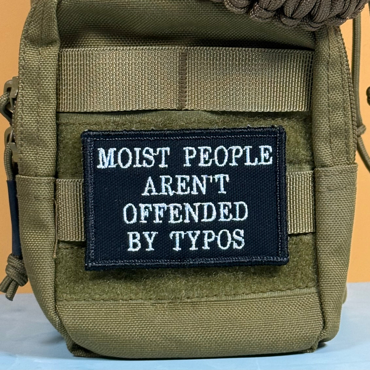 Moist People Aren’t Offended by Typos embroidered morale patch: olive green tactical pouch with a black embroidered patch that reads 'MOIST PEOPLE AREN'T OFFENDED BY TYPOS' in white text, shown against a solid orange background.