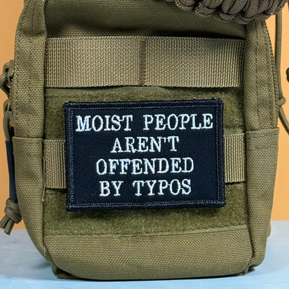 Moist People Aren’t Offended by Typos embroidered morale patch: olive green tactical pouch with a black embroidered patch that reads 'MOIST PEOPLE AREN'T OFFENDED BY TYPOS' in white text, shown against a solid orange background.