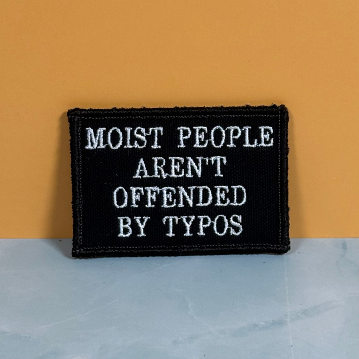 Moist People Aren’t Offended by Typos embroidered morale patch: black embroidered patch with white text reading 'MOIST PEOPLE AREN'T OFFENDED BY TYPOS', photographed against a mustard-yellow wall and pale blue-gray surface.