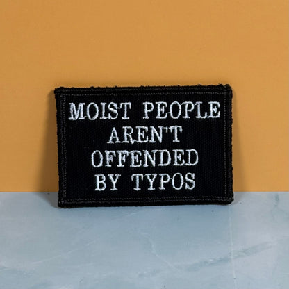 Moist People Aren’t Offended by Typos embroidered morale patch: black embroidered patch with white text reading 'MOIST PEOPLE AREN'T OFFENDED BY TYPOS', photographed against a mustard-yellow wall and pale blue-gray surface.