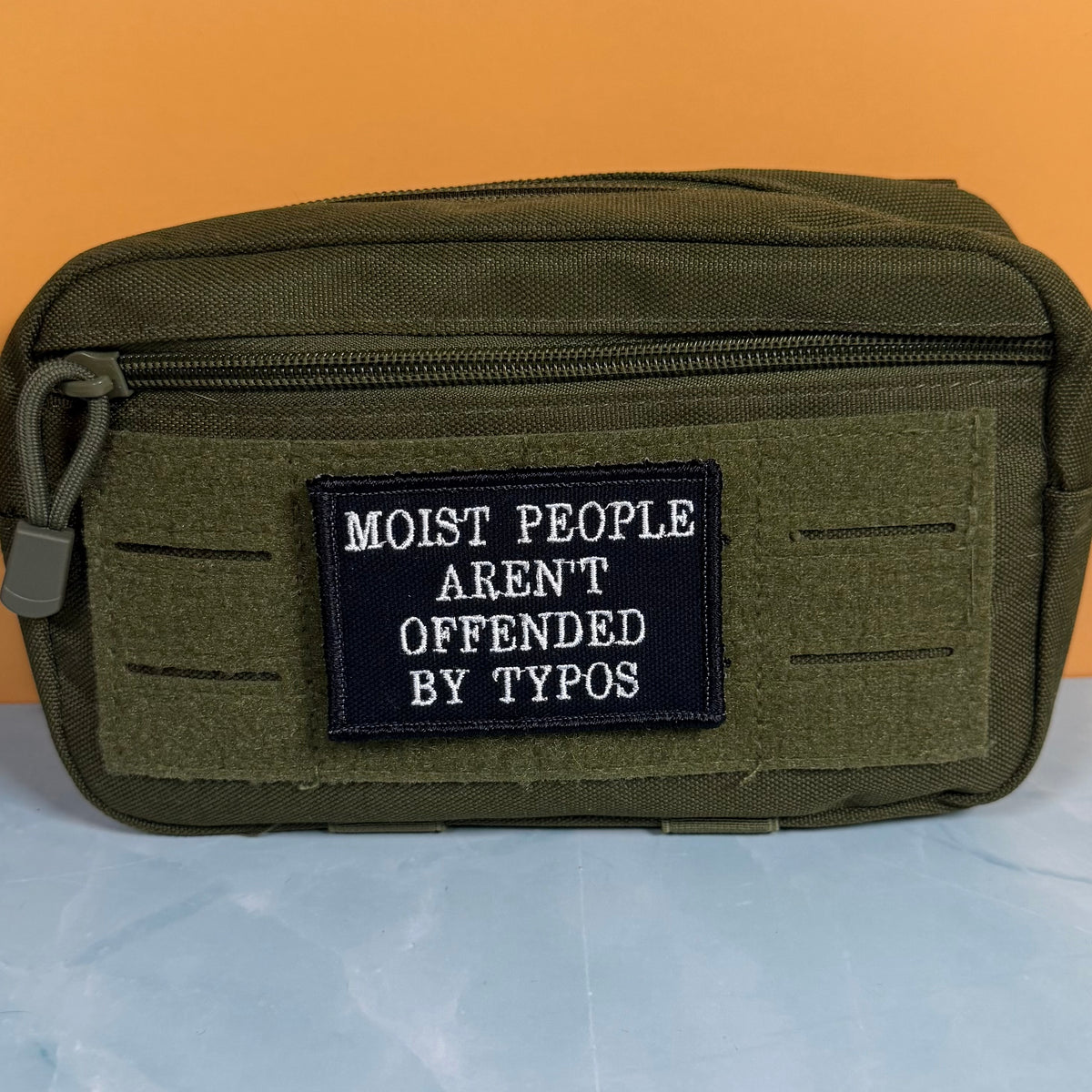 Moist People Aren’t Offended by Typos embroidered morale patch: olive green tactical pouch with a black embroidered patch that reads 'MOIST PEOPLE AREN'T OFFENDED BY TYPOS' in white text, set against a split orange and light.