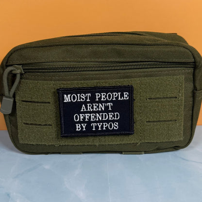 Moist People Aren’t Offended by Typos embroidered morale patch: olive green tactical pouch with a black embroidered patch that reads 'MOIST PEOPLE AREN'T OFFENDED BY TYPOS' in white text, set against a split orange and light.
