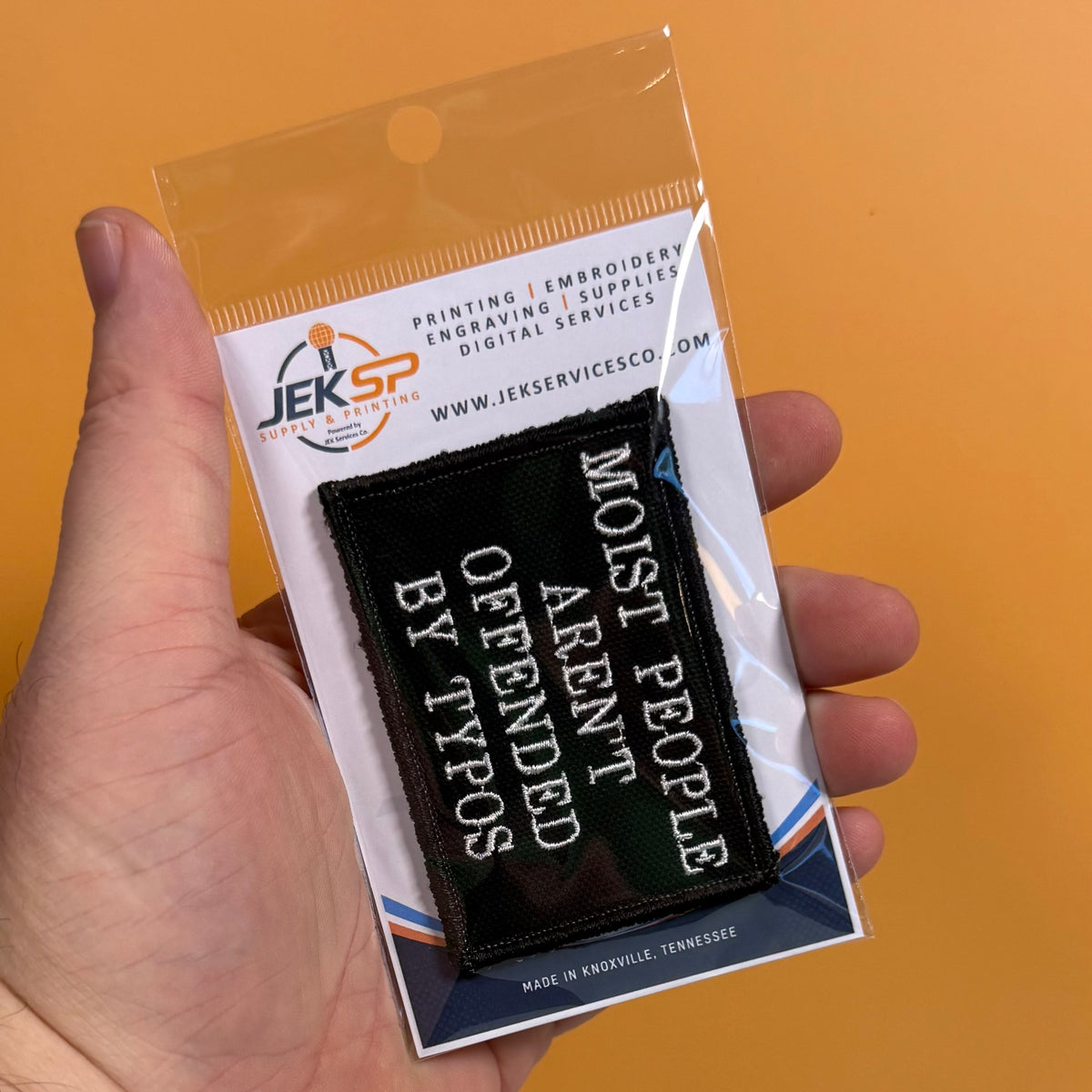 Moist People Aren’t Offended by Typos embroidered morale patch: hand holds a clear plastic package containing a black embroidered patch with white text reading 'MOST PEOPLE ARE N'T OFFENDED BY TYPOS.' The package card shows.