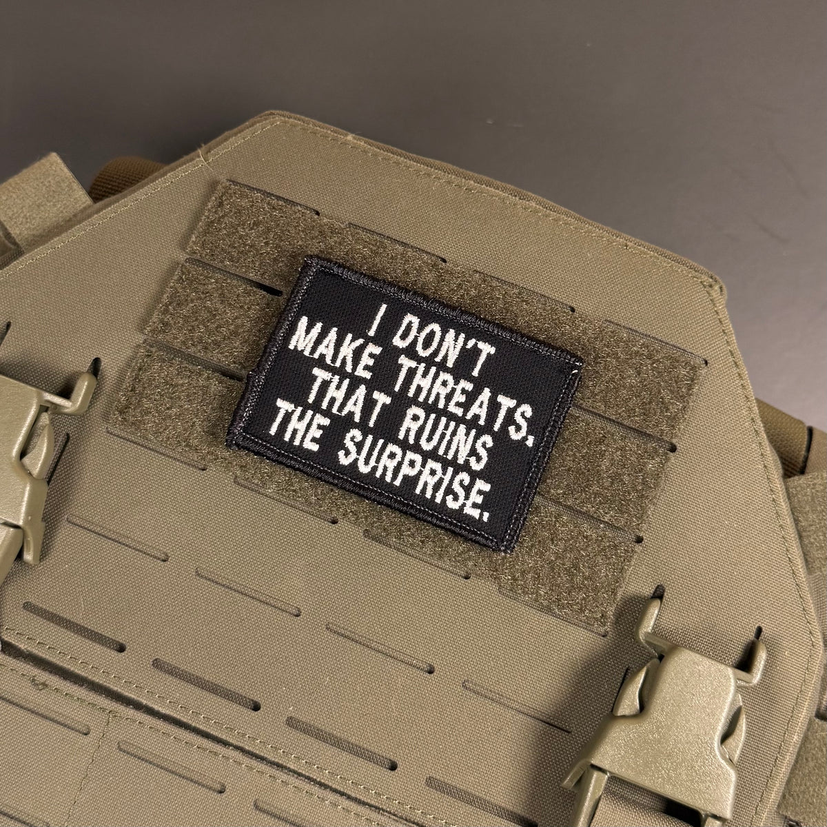 A rectangular black tactical patch with white embroidered text reading 'I DON'T MAKE THREATS. THAT RUINS THE SURPRISE.