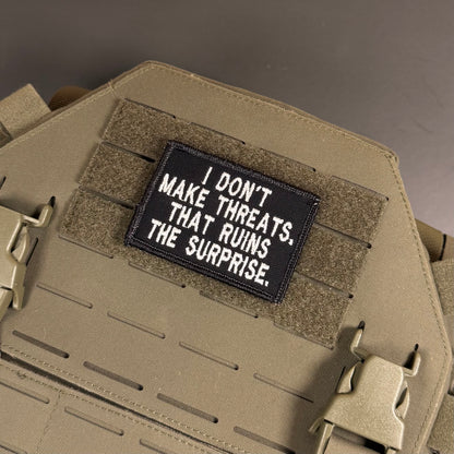 A rectangular black tactical patch with white embroidered text reading 'I DON'T MAKE THREATS. THAT RUINS THE SURPRISE.