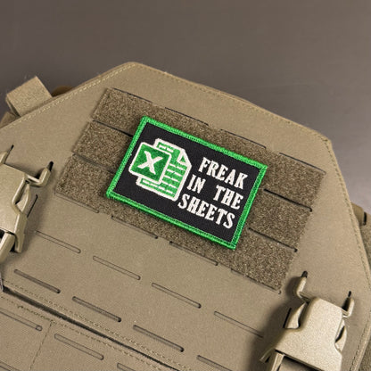 A rectangular embroidered tactical patch with a black background, green border, Excel-style green spreadsheet icon, and the words 'FREAK IN THE SHEETS' in white, mounted on tan MOLLE webbing.