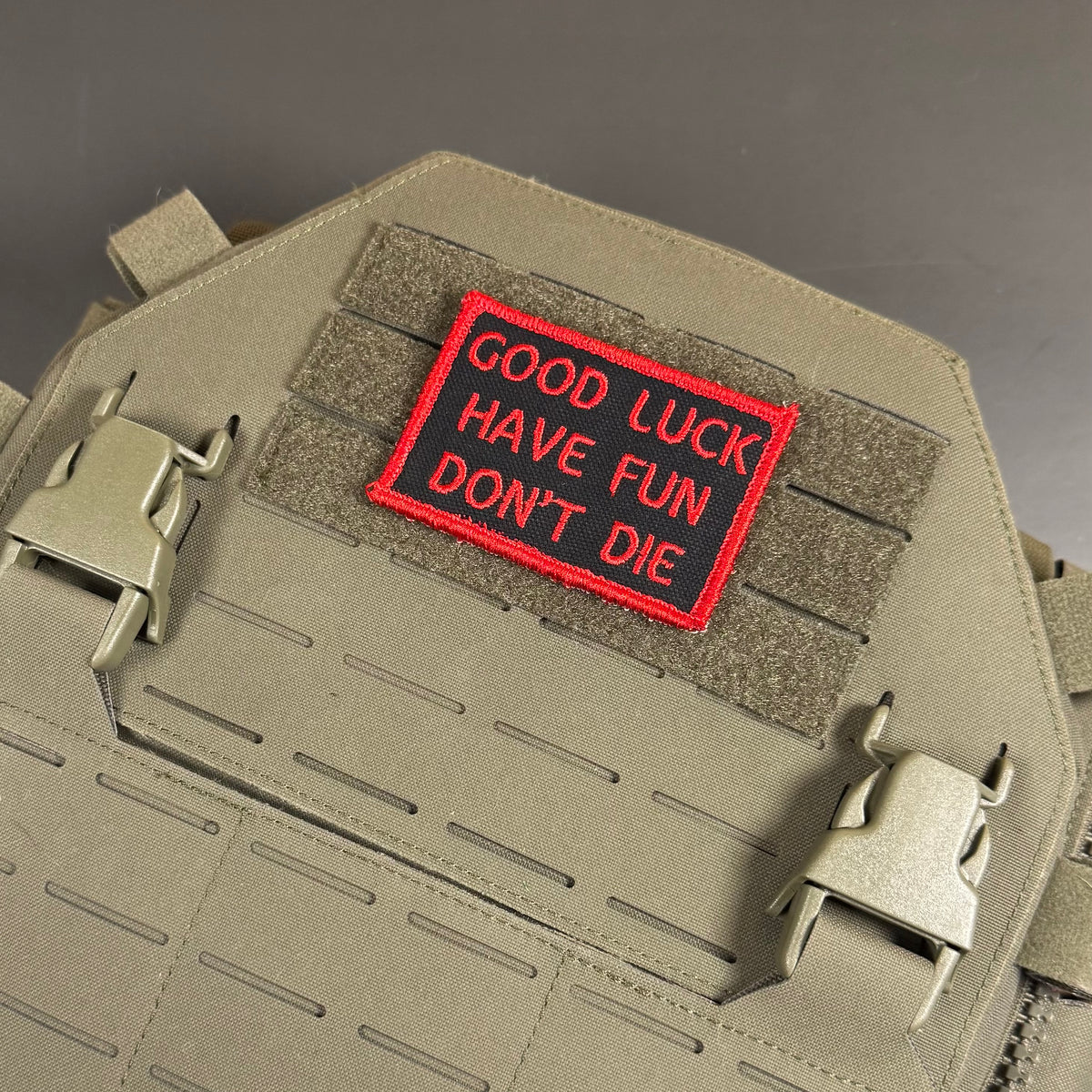 A rectangular embroidered patch with red lettering reading 'GOOD LUCK HAVE FUN DON'T DIE' on a red background, attached to olive drab tactical gear with velcro and metal buckles.