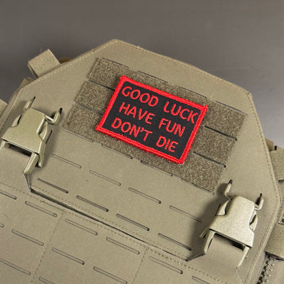 A rectangular embroidered patch with red lettering reading 'GOOD LUCK HAVE FUN DON'T DIE' on a red background, attached to olive drab tactical gear with velcro and metal buckles.