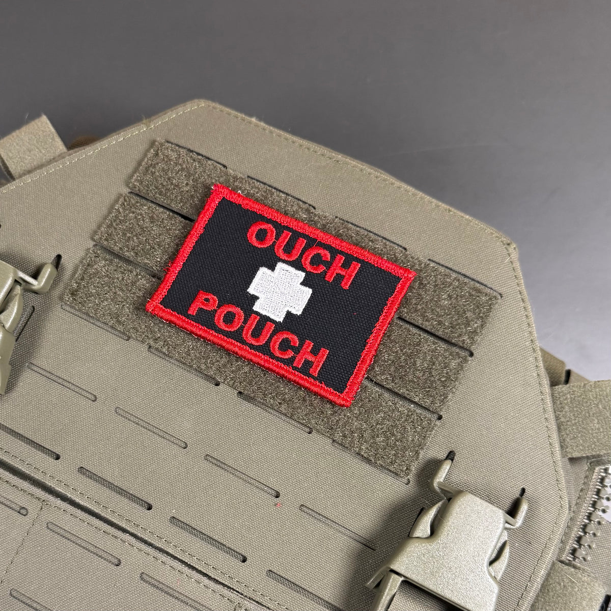An embroidered tactical patch with the text 'OUCH POUCH' in red on a black background with a white cross, attached to a tan MOLLE webbing panel on a matching tan tactical vest or backpack.