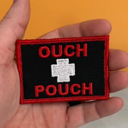 A rectangular embroidered patch held in a hand, featuring a black background with red border, red text reading 'OUCH POUCH,' and a white cross in the center.