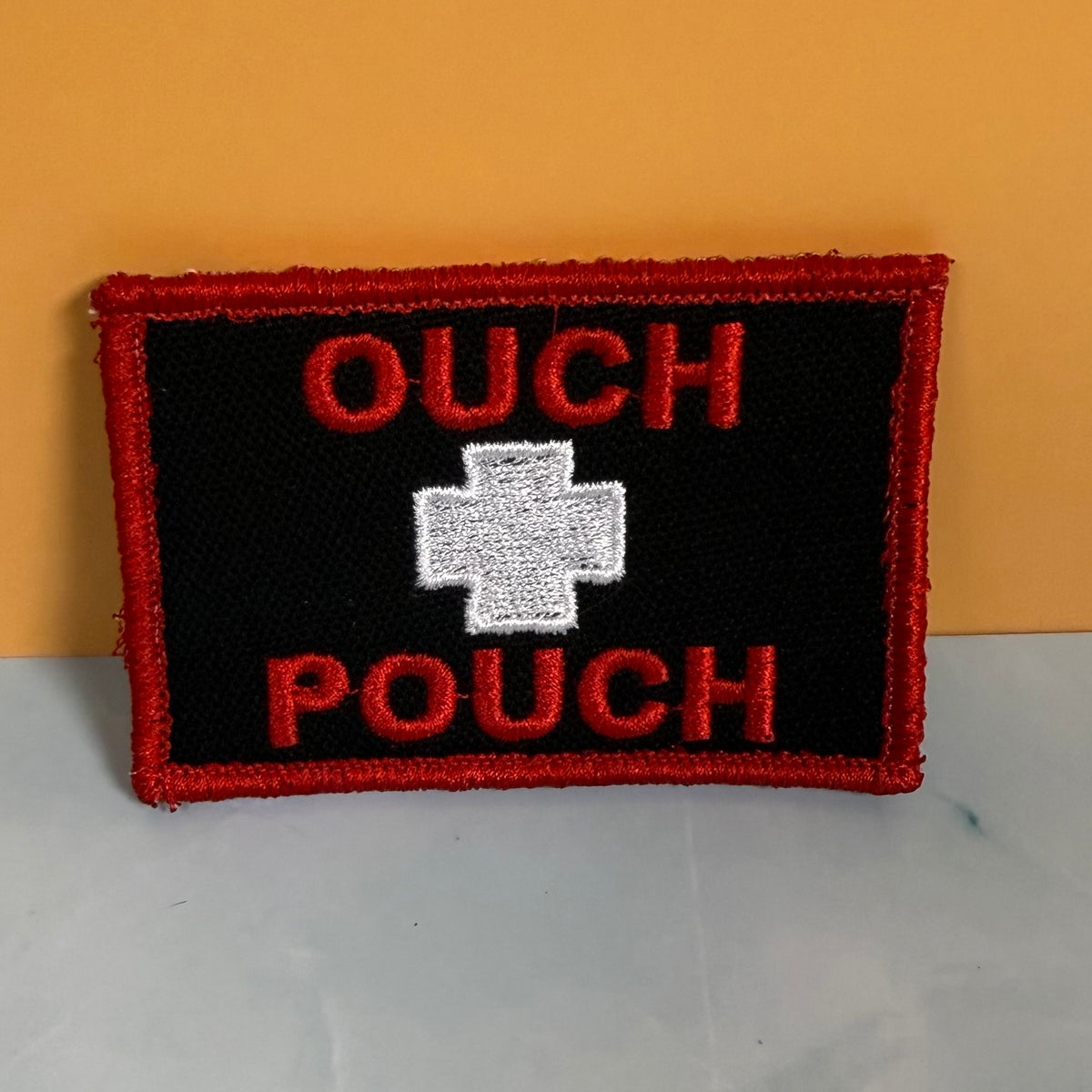 An embroidered patch with a black background, red border, red text reading 'OUCH POUCH', and a white cross in the center, photographed against a yellow wall and light surface.