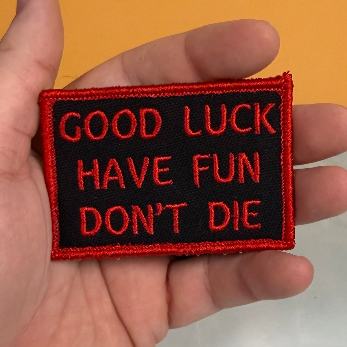 A rectangular embroidered patch held in a hand, featuring the text 'GOOD LUCK HAVE FUN DON'T DIE' in red embroidery on a black background with a red border.