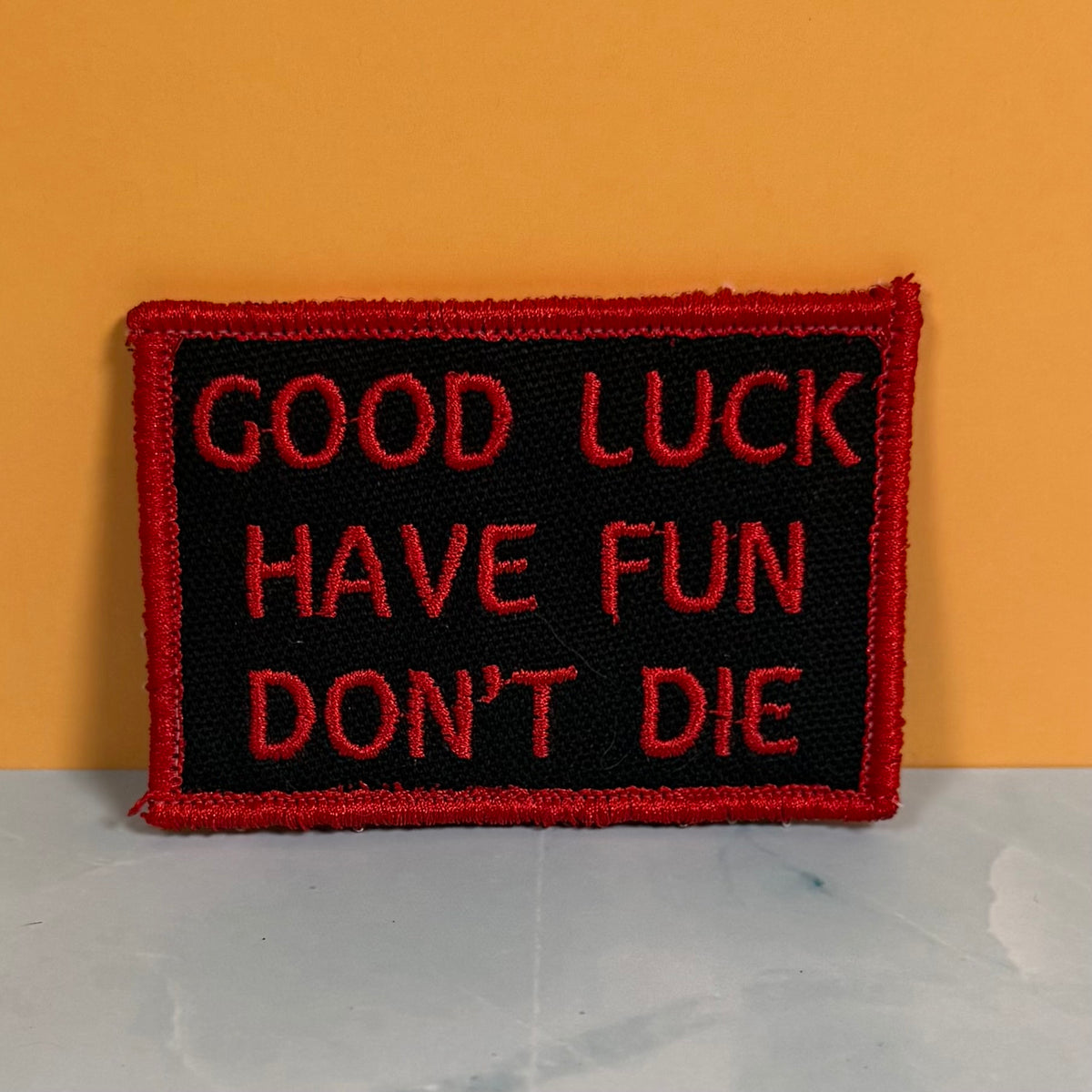 A rectangular embroidered patch with a black center, red border, and red text reading 'GOOD LUCK HAVE FUN DON'T DIE', photographed against a warm orange background and light gray surface.