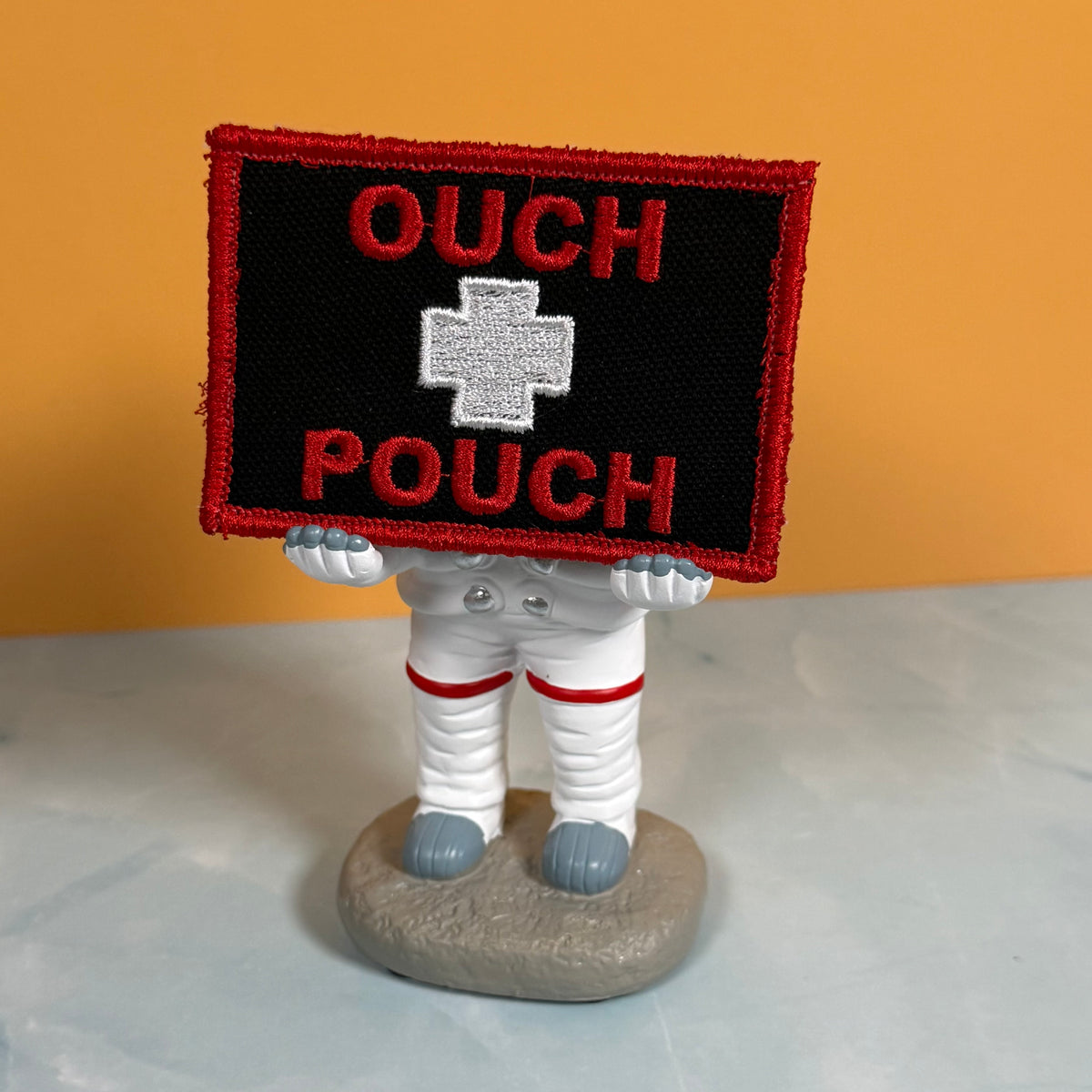 An embroidered patch with a red border, black background, red 'OUCH' text at the top and bottom, and a white cross in the center, held by a small astronaut figurine on a gray base against an orange and white background.