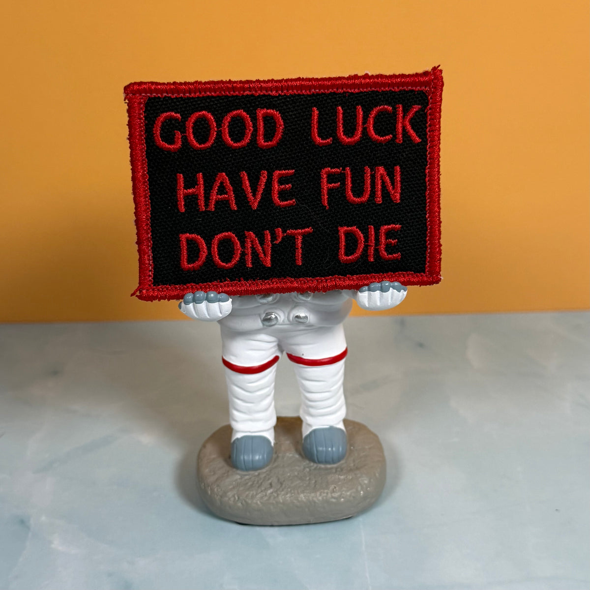 An embroidered patch with the text 'GOOD LUCK HAVE FUN DON'T DIE' in red on a black background with a red border, held by a small astronaut figurine on a gray base against an orange and gray background.