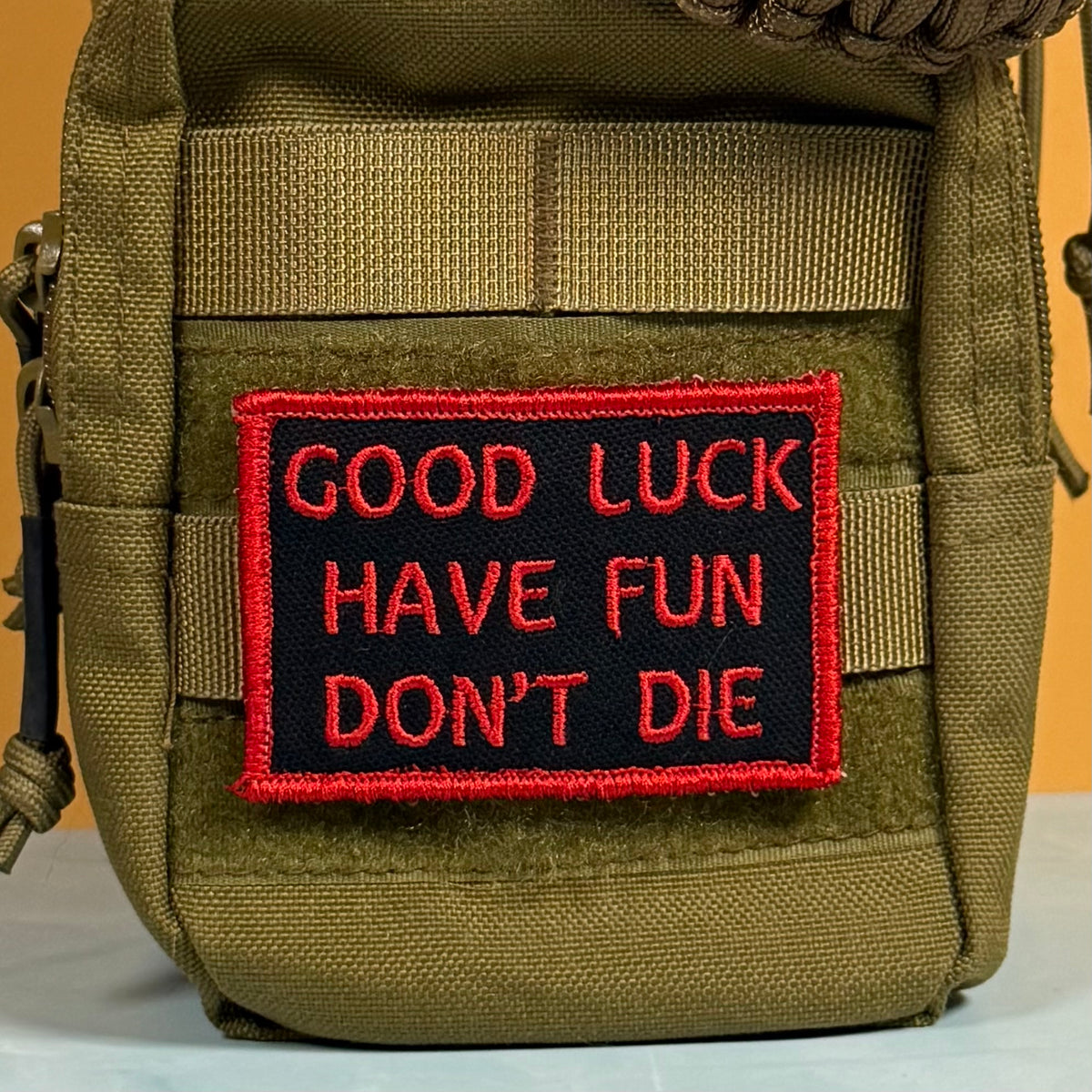 A tactical utility pouch in olive drab green nylon with a black rectangular embroidered patch featuring red text that reads 'GOOD LUCK HAVE FUN DON'T DIE' and a red border.