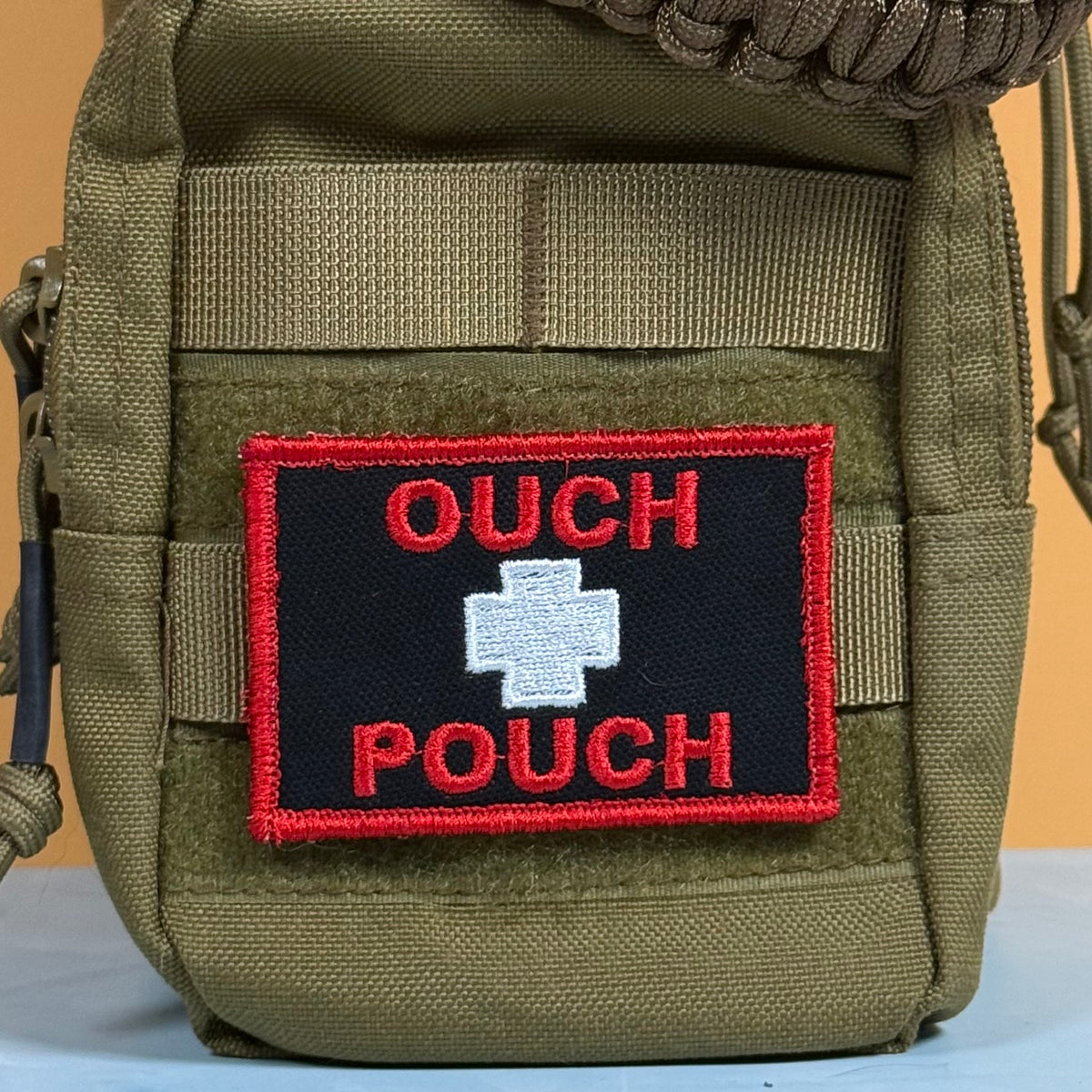 An olive green tactical pouch featuring a black rectangular embroidered patch with a white cross in the center and the words 'OUCH POUCH' in red.
