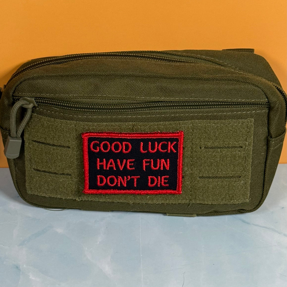 An olive green tactical pouch with a front zipper and a red embroidered patch that reads 'GOOD LUCK HAVE FUN DON'T DIE' on a black background, set against an orange and light blue background.