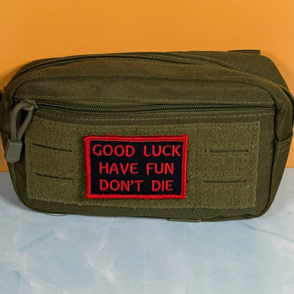 An olive green tactical pouch with a front zipper and a red embroidered patch that reads 'GOOD LUCK HAVE FUN DON'T DIE' on a black background, set against an orange and light blue background.