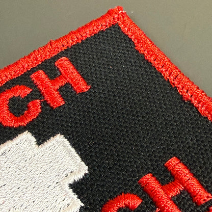 Close-up of an embroidered patch showing a black background with red border and red lettering, plus a white graphic element, photographed on a neutral gray surface.