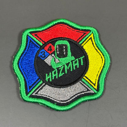 An embroidered tactical patch featuring a Maltese cross design with a green border, red, blue, yellow, and gray sections, and a central black circle with a green construction helmet and the text 'HAZMAT'.