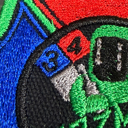 Close-up of a colorful embroidered patch featuring a green alien head with black eyes, a red square with the number 4, a blue square with the number 3, and a black circular border.