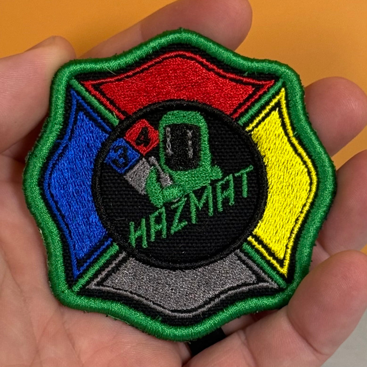 An embroidered Hazmat patch held in a person's hand. The patch features a Maltese cross design with red, blue, yellow, green, and gray sections surrounding a central black circle with a green hazmat suit and the word 'HAZMAT'.