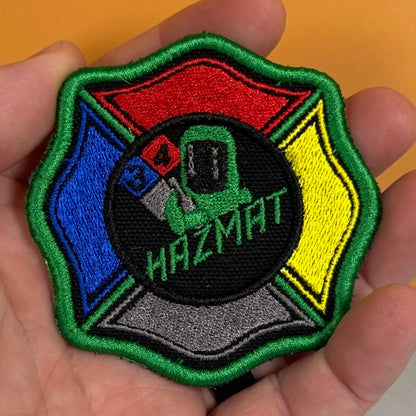 An embroidered Hazmat patch held in a person's hand. The patch features a Maltese cross design with red, blue, yellow, green, and gray sections surrounding a central black circle with a green hazmat suit and the word 'HAZMAT'.