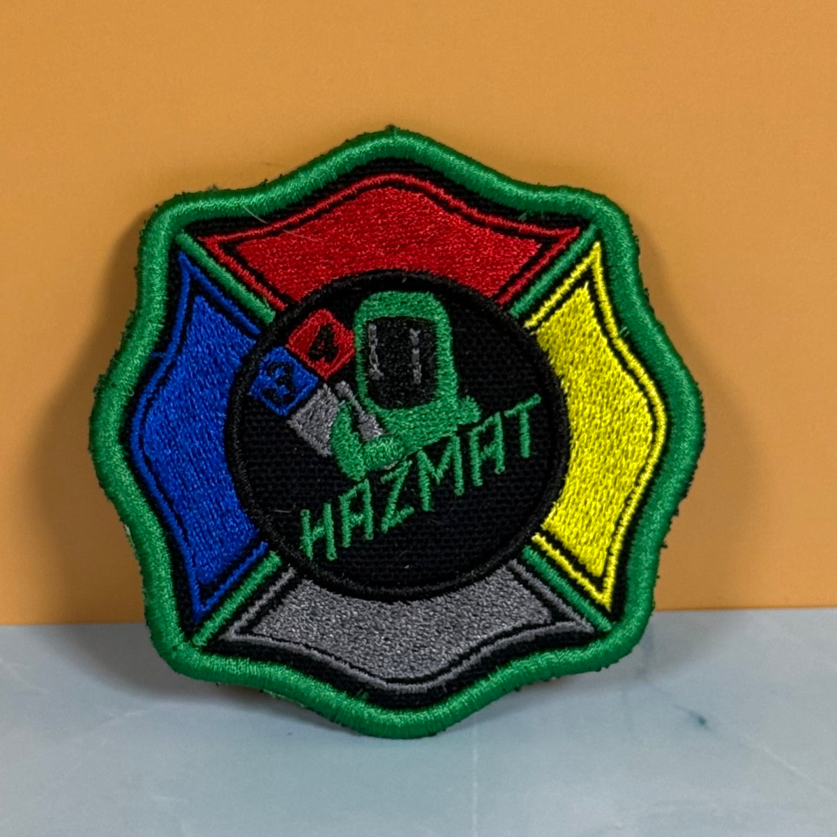 An embroidered Hazmat patch shaped like a Maltese cross, featuring a green border, red, blue, yellow, and gray sections surrounding a central black circle with a green hazmat suit and the word 'HAZMAT'.