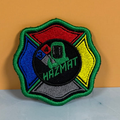 An embroidered Hazmat patch shaped like a Maltese cross, featuring a green border, red, blue, yellow, and gray sections surrounding a central black circle with a green hazmat suit and the word 'HAZMAT'.