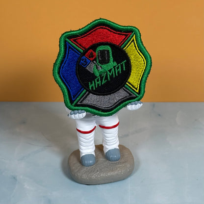 An embroidered HAZMAT firefighter patch displayed on a small astronaut figurine standing on a gray base, set against a two-tone orange and gray background.