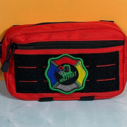 A red zippered utility pouch with a black velcro panel on the front featuring a colorful Hazmat patch, set against a solid orange background.