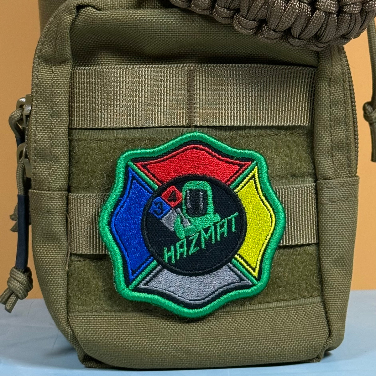 An olive drab tactical pouch featuring a colorful embroidered Hazmat patch on the front, with a paracord strap visible at the top.